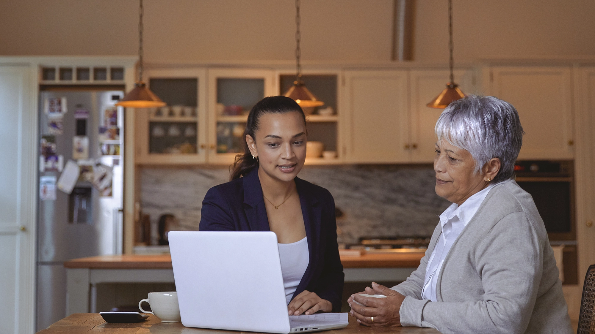 A senior woman receives help with her finances in the comfort of her cozy kitchen at home. The kitchen, warmly lit and inviting, features charming details like a wooden table and family photos on the fridge. Seated at the table, the woman reviews financial documents with a caring advisor or family member, who patiently explains details and provides assistance.